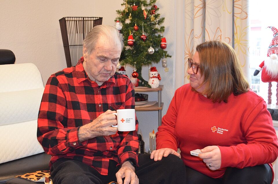 Älterer Herr mit Tasse sitzt auf Sofa, daneben eine Pflegefachkraft mit Tabletten