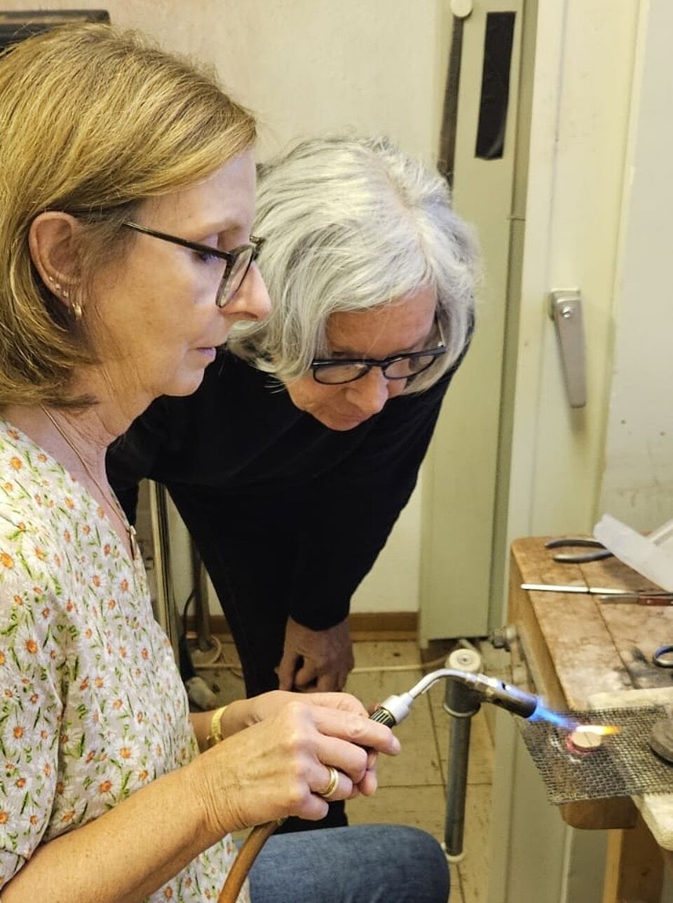 Kursleiterin unterstützt beim Schmuckerstellen Eine Frau lötet ein neues Schmuckstück, während ihr über die Schulter geschaut wird