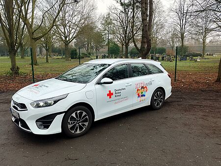 Zu sehen ist ein weißes Auto von der Seite, mit der Aufschrift ,,Kita Flitzer" und dem Logo des DRK Duisburg. Es handelt sich hierbei um den neuen Kita Flitzer